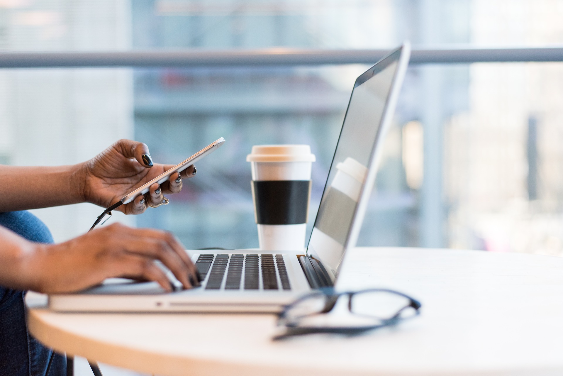 A person working at a table with a laptop and smartphone. One hand is typing on the laptop keyboard, while the other holds a smartphone connected via a charging cable. A takeaway coffee cup and a pair of glasses are placed next to the laptop on the table. The background is blurred, suggesting a casual, modern workspace or cafe setting.