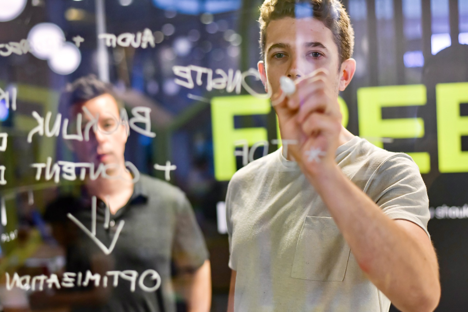 Two men working together in an office setting, writing on a transparent glass board. One person is writing with a marker, while the other is observing the notes. The writing on the board includes terms like 'Backlink,' 'Content,' and 'Optimization,' suggesting a brainstorming session or strategy planning related to SEO or digital marketing. The background is slightly blurred, with a large, bright green sign partially visible.