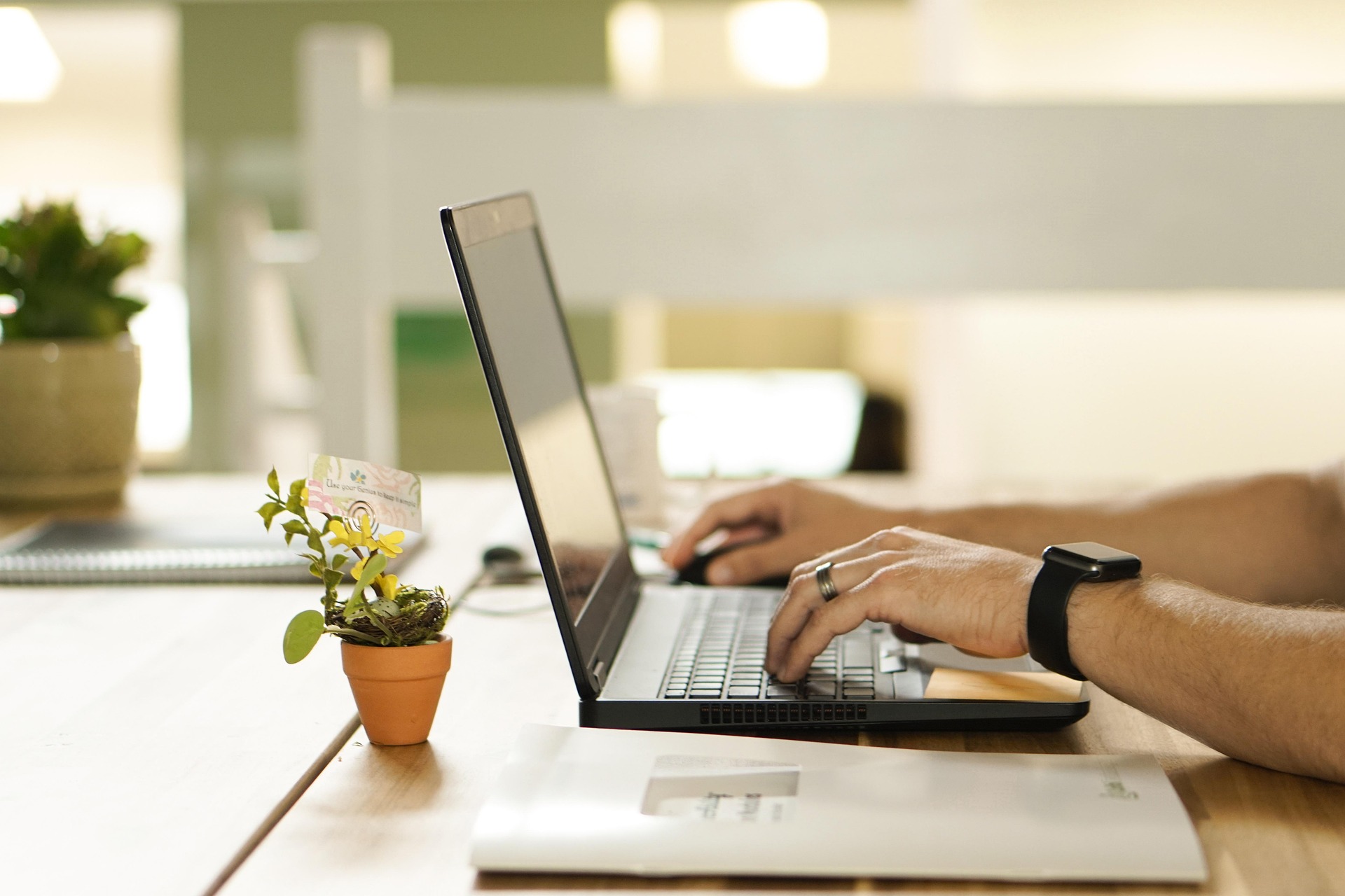 A person working on a laptop at a wooden desk, typing with both hands. The individual is wearing a smartwatch and a wedding ring. On the desk, there is a small potted plant with yellow flowers and a decorative card, alongside a notebook and an envelope. The background is softly blurred, creating a calm and focused work environment, possibly in a home office or co-working space.