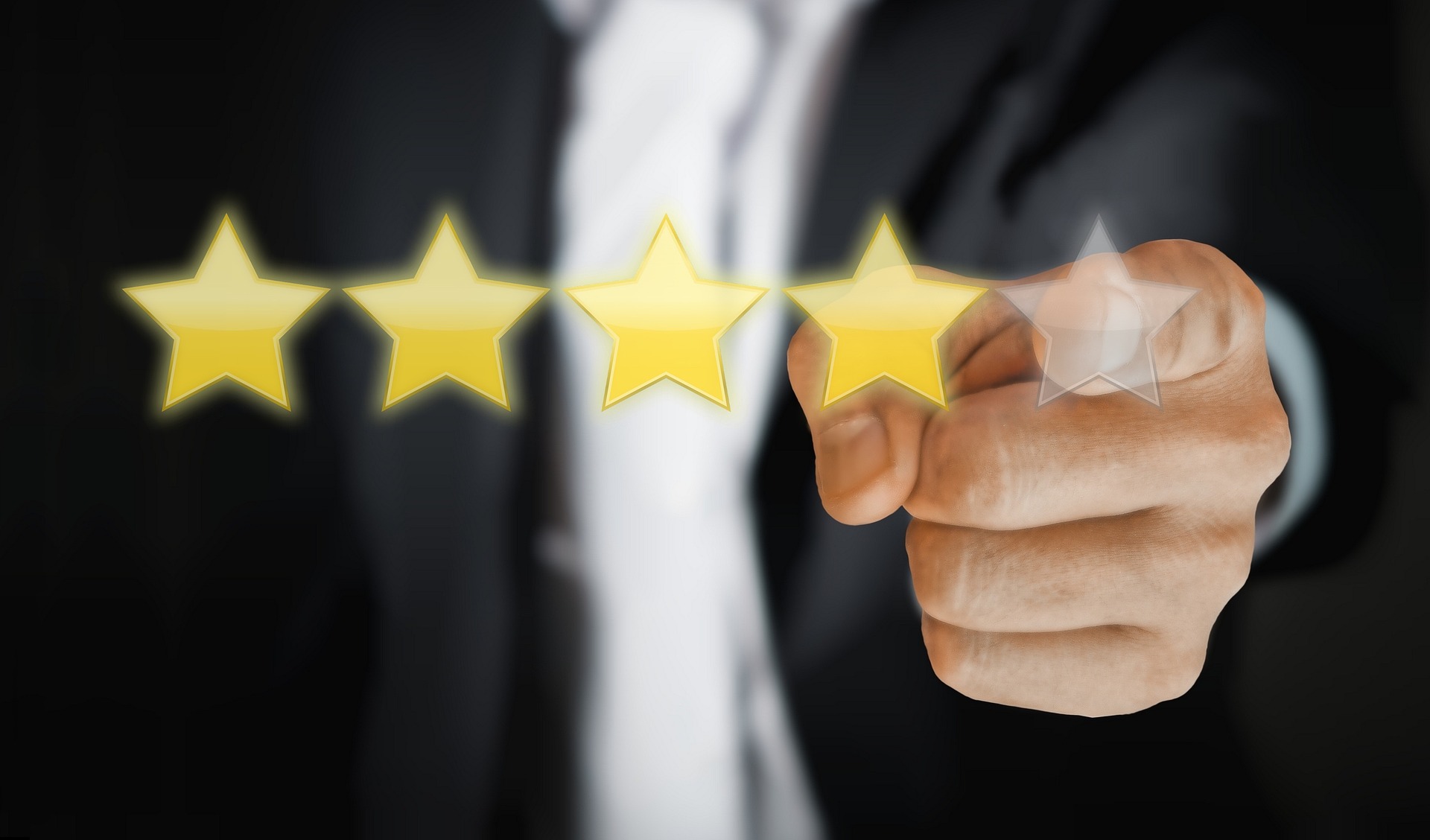 A close-up image of a hand pointing towards a row of five stars, with four of the stars glowing yellow and the fifth one partially transparent. The person is wearing a business suit, and the gesture represents a rating or review system, commonly used in customer feedback or product reviews.