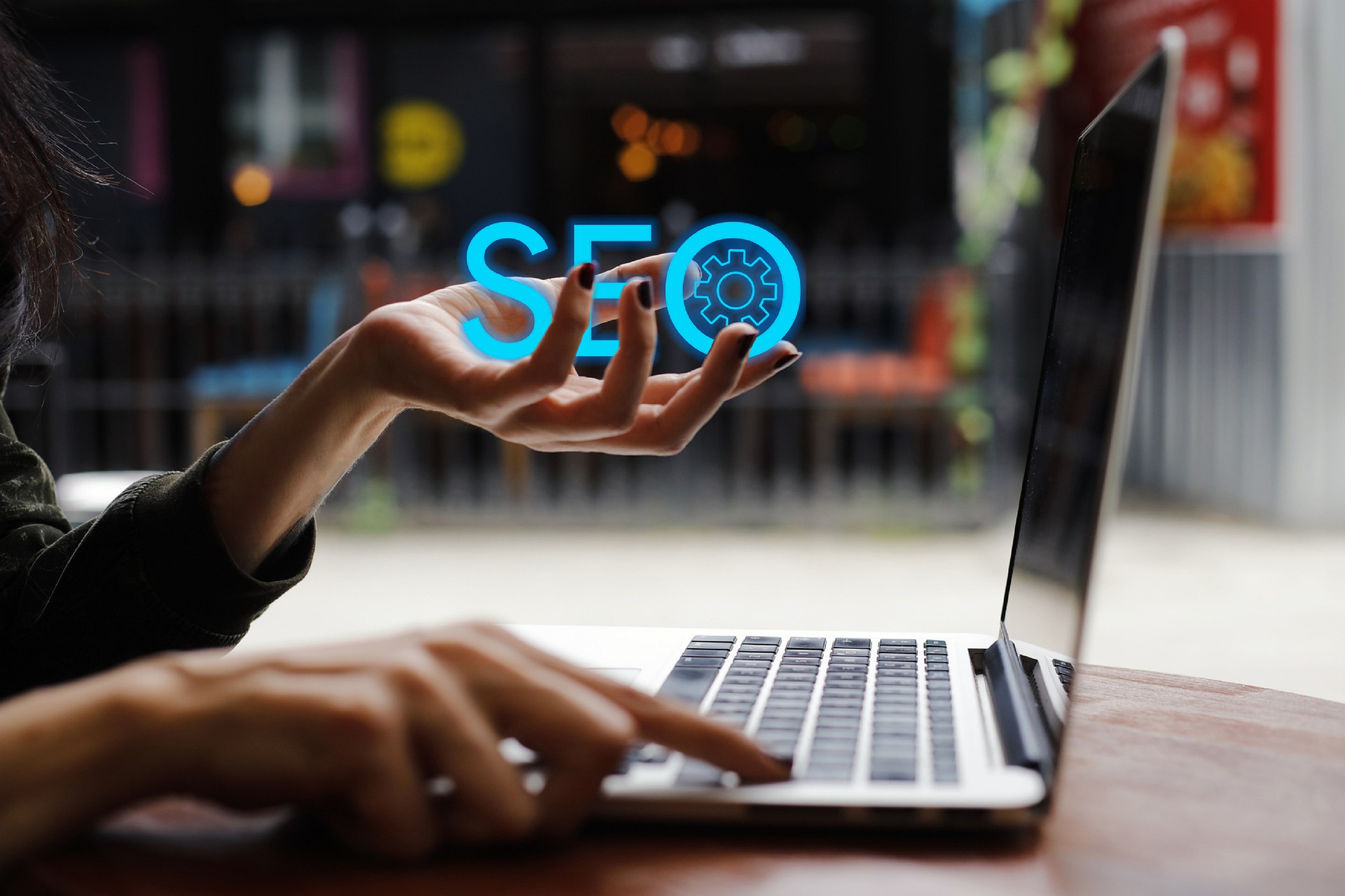 A person working on a laptop with one hand typing on the keyboard and the other hand holding a glowing blue 'SEO' icon with a gear symbol. The background is blurred, focusing attention on the hand and the laptop. The image represents SEO (Search Engine Optimization) and its technical aspects, symbolized by the gear, in the context of digital marketing or website management.