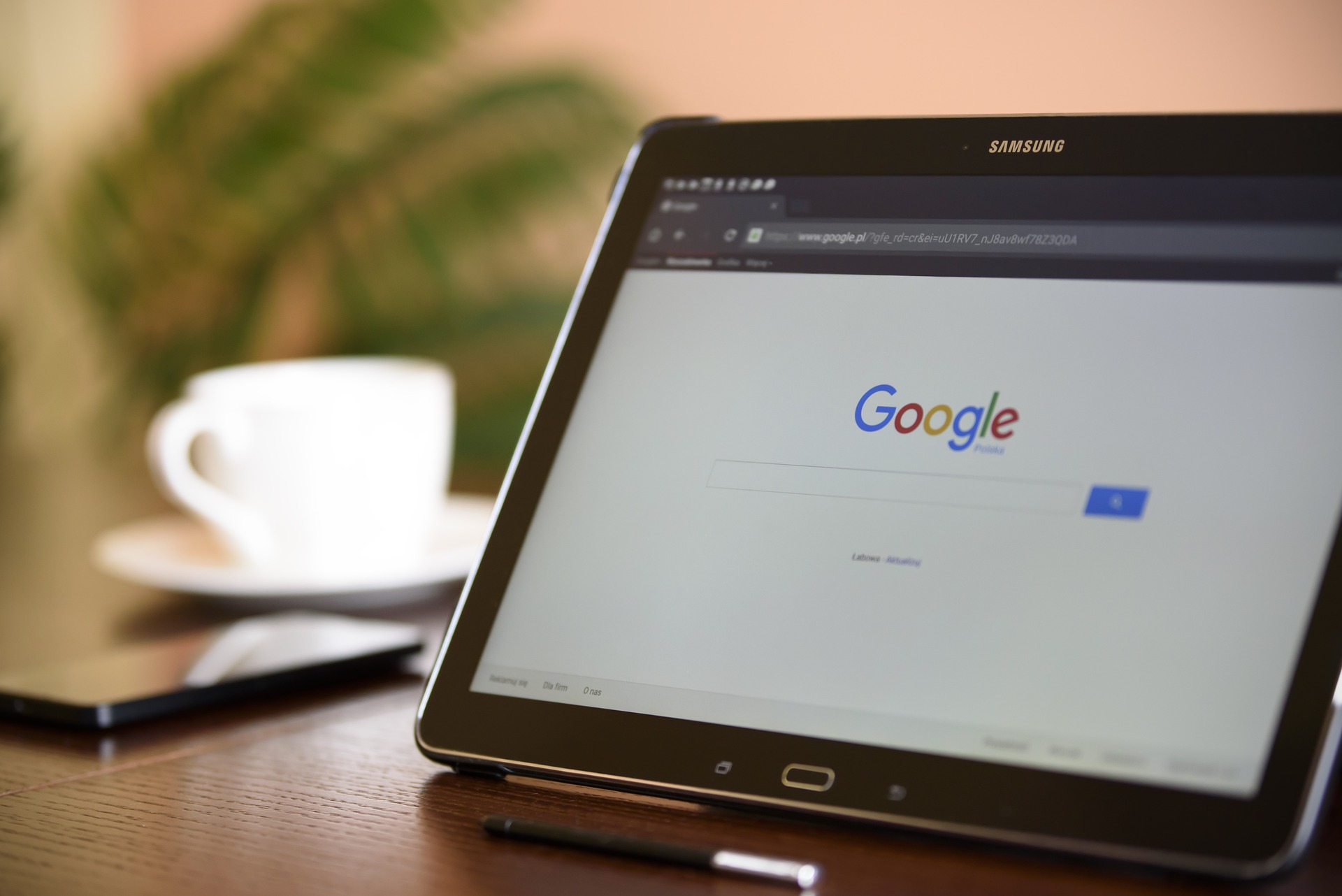 A Samsung tablet displaying the Google search homepage is placed on a wooden desk. Next to the tablet, there is a smartphone and a stylus pen. In the background, a blurred coffee cup sits on a saucer. The image suggests a work or research setup, with a focus on online browsing or SEO activities.