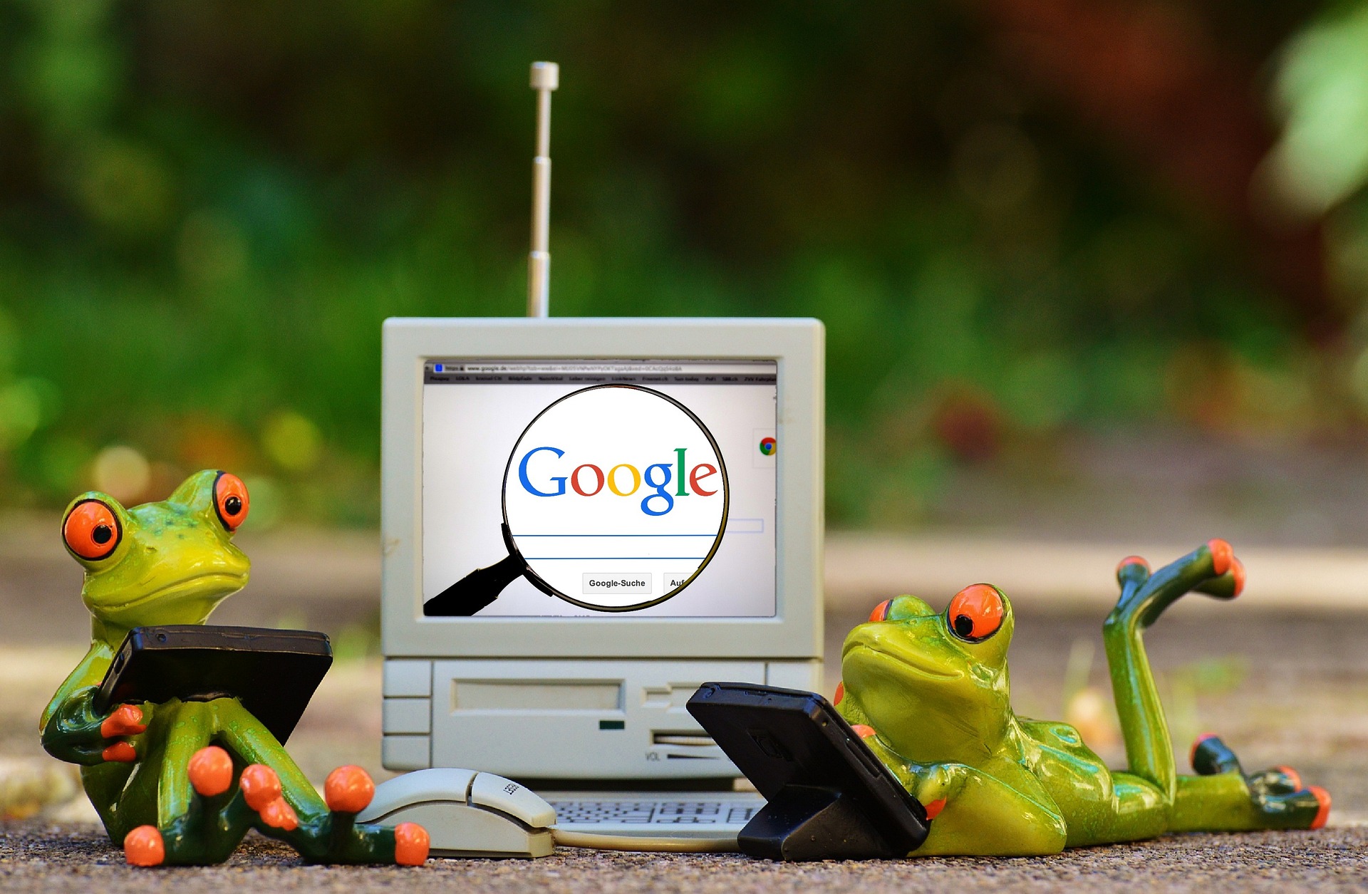 A playful scene featuring two cartoonish frog figurines using tablets while sitting and lying beside an old-school computer. The computer screen displays the Google homepage with a magnifying glass highlighting the Google logo. One frog is seated upright holding a tablet, while the other frog is reclining, also holding a tablet. The background is an outdoor, blurred natural setting, adding to the whimsical nature of the image. The scene humorously represents technology and search engine usage.