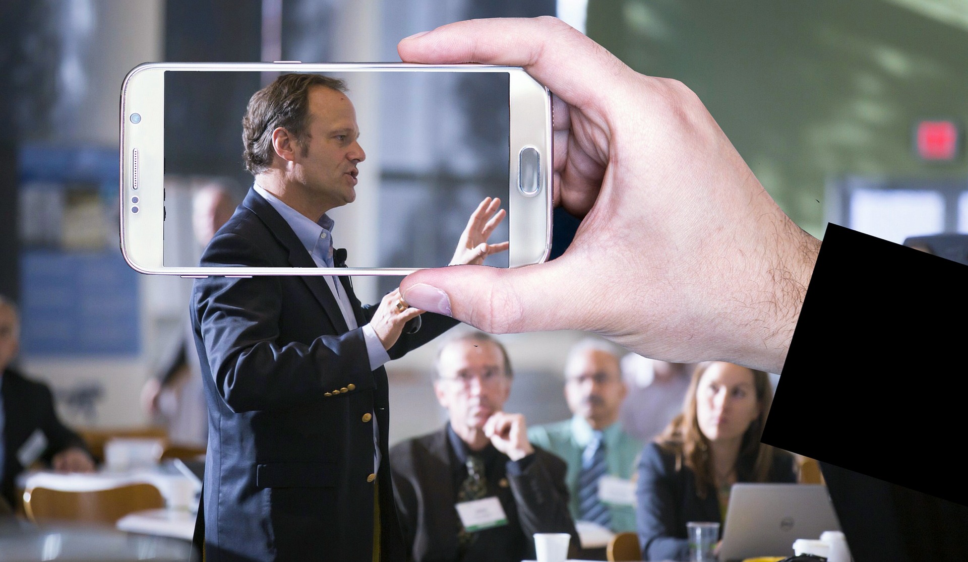 This image shows a man giving a speech or presentation in a formal setting, captured through the lens of a smartphone. The hand of the person holding the phone frames the speaker, who is gesturing with one hand as he talks. The background features an audience, some of whom are listening attentively, with a blurred office or conference setting. The image emphasizes modern communication, where moments are often captured and shared via mobile devices.