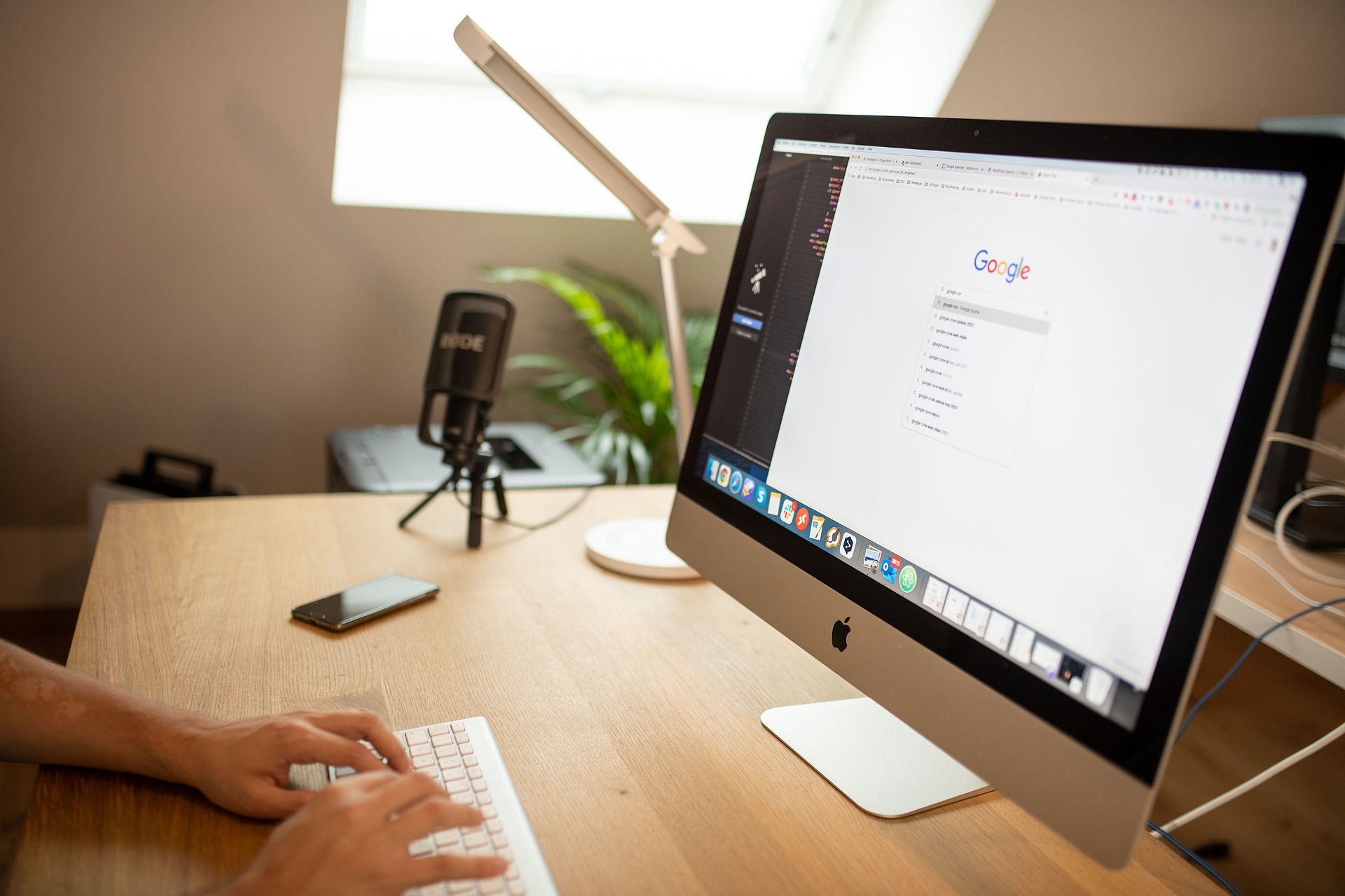 A person working at a desk, typing on an Apple keyboard in front of an iMac displaying the Google search homepage. On the desk, there is a microphone (labeled 'RØDE'), a smartphone, and a desk lamp. The scene suggests a workspace setup for tasks like SEO, content creation, or digital marketing, with a focus on online research and copywriting.