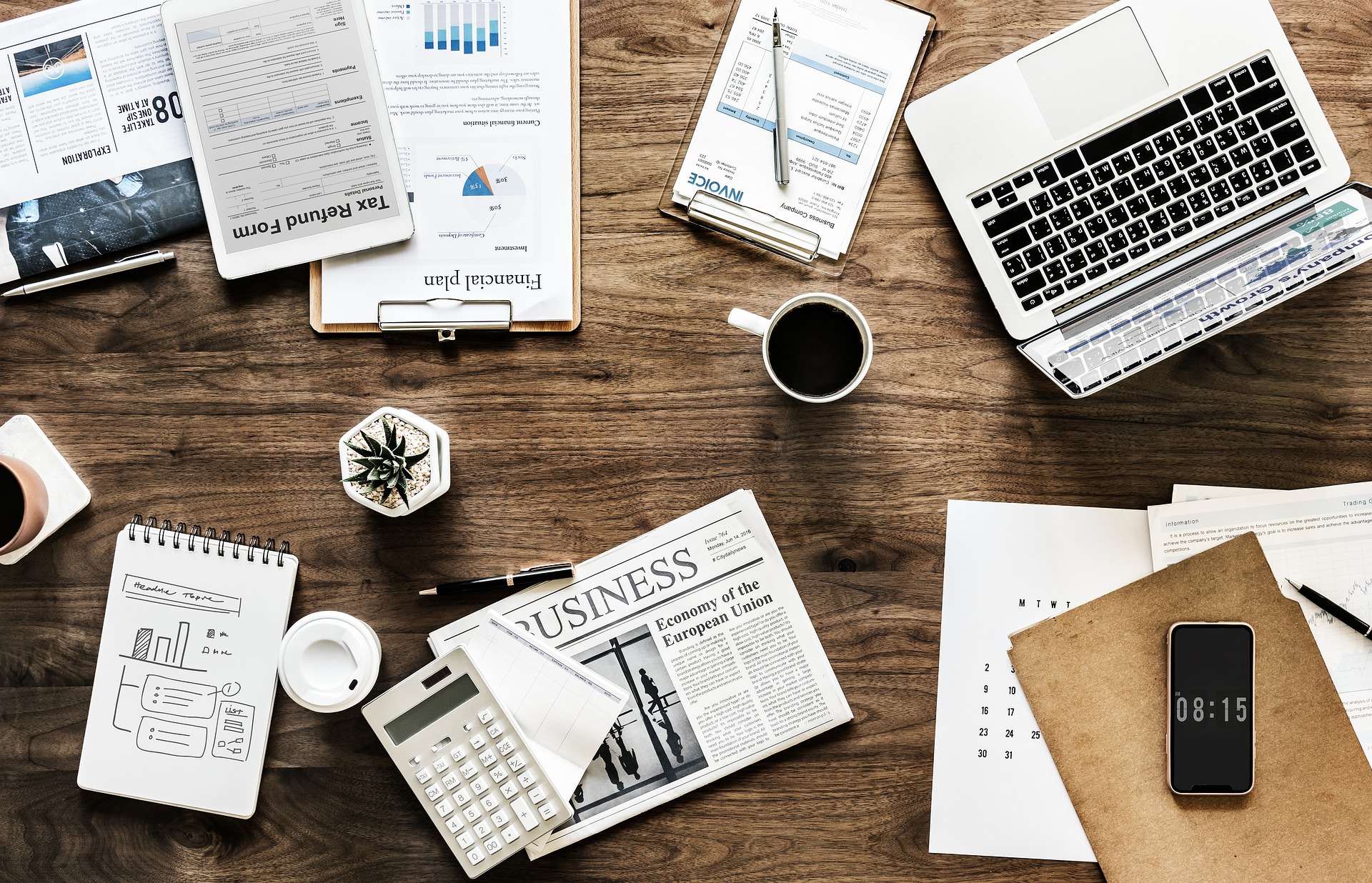 A wooden desk covered with various business-related items. On the desk are a laptop, calculator, notebook with sketches, invoices, financial documents, a tax refund form, and a newspaper with the headline 'Economy of the European Union.' There are also two cups of coffee, a smartphone displaying the time as 08:15, a small potted succulent plant, and a clipboard. The scene conveys a busy, organized workspace focused on financial planning and business activities.