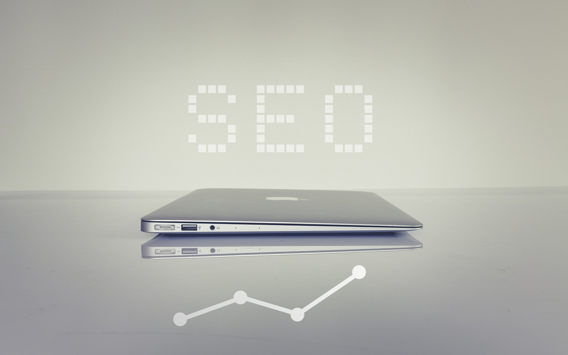 A minimalist image showing a closed laptop lying flat on a reflective surface. Above the laptop, the word 'SEO' is displayed in a pixelated font. Below the laptop, a line graph with dots indicates an upward trend. The image represents the concept of SEO (Search Engine Optimization) and its role in improving website rankings.