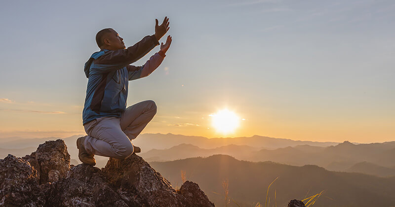 Man kneeling on a mountain, outstretching arms up in the air.